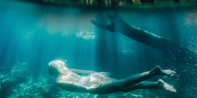 An underwater scene of a woman in a one piece white bathing suit swimming gently through turquoise water, past a submerged branch, as shafts of sunlight dance over her body. 